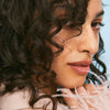 Eine Frau mit lockigem Haar und natürlichem Make-up blickt zur Seite und präsentiert Ergebnis des Coils & Curls Weightless Curl Definer von Maria Nila.