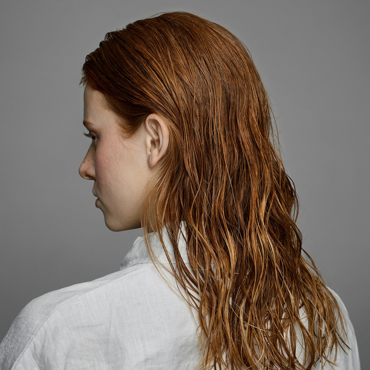 Eine Person mit nassem, schulterlangem rotem Haar vor einem grauen Hintergrund -zeigt die leuchtende Wirkung des maria nila Luminous Colour Shampoo.