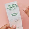 Eine Hand hält eine aufgerissene Packung Mario Badescu Drying Patch und zeigt das transparente Pflasterblatt darin.