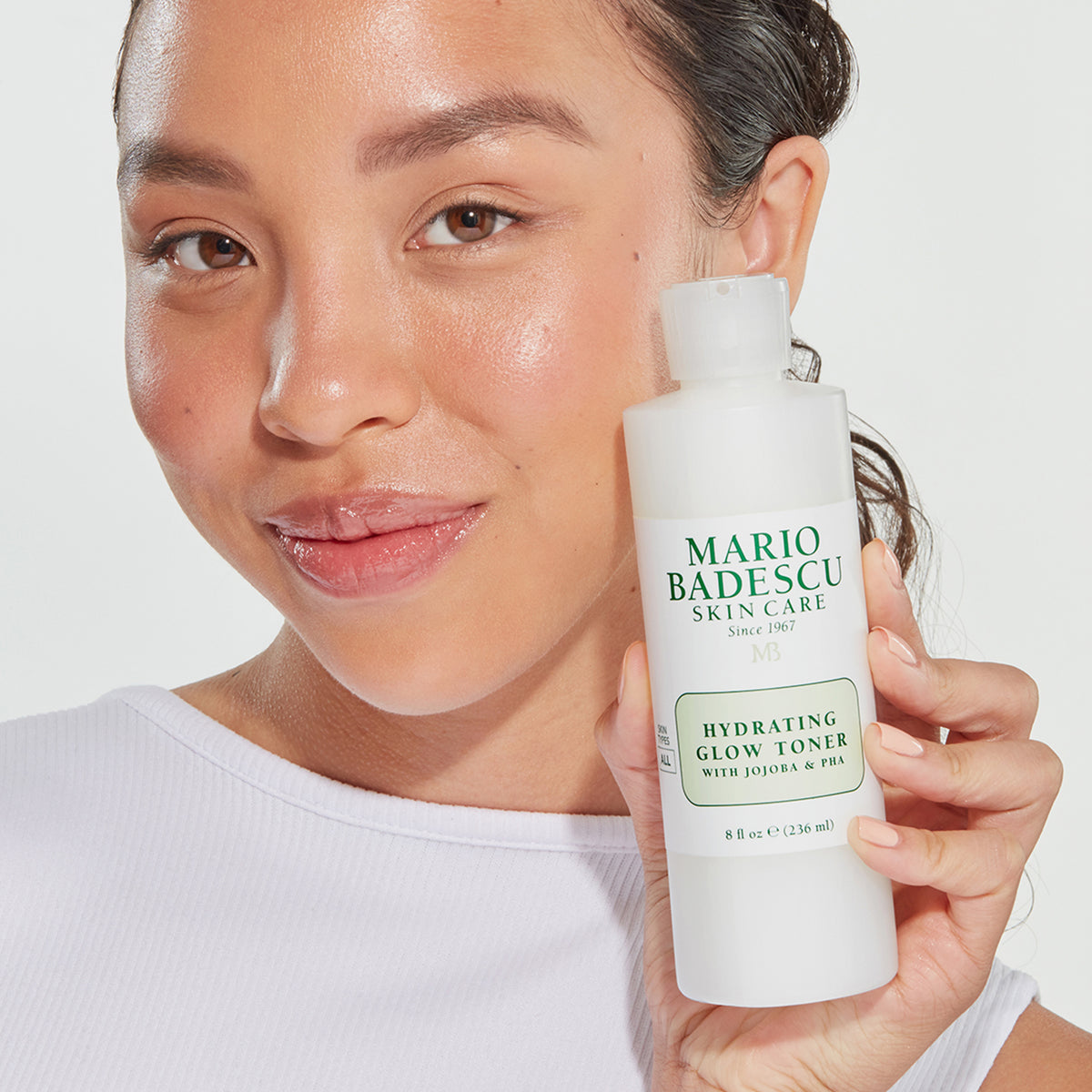 Eine lächelnde Frau hält den Mario Badescu Hydrating Glow Toner in der Hand und zeigt damit strahlende, leuchtende Haut.