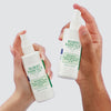 Zwei Hände halten Mario Badescu Repairing Facial Spray with Hypochlorous Acid Flaschen vor einem hellgrauen Hintergrund.