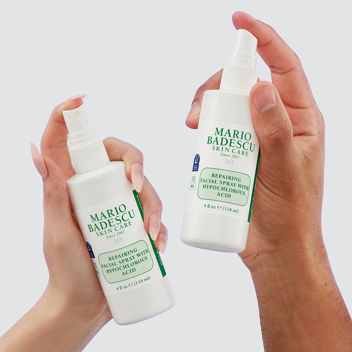 Zwei Hände halten Mario Badescu Repairing Facial Spray with Hypochlorous Acid Flaschen vor einem hellgrauen Hintergrund.