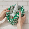Hände, die eine grün karierte Tasche mit Mario Badescu-Produkten, darunter das Repairing Facial Spray with Hypochlorous Acid, auf einer weiß gekachelten Fläche halten.