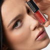 Eine Frau hält einen Mesauda Beauty LIPOILOGY™ Lipgloss in die Nähe ihres Auges, um ihre glänzenden Lippen zu betonen und blickt in die Kamera.