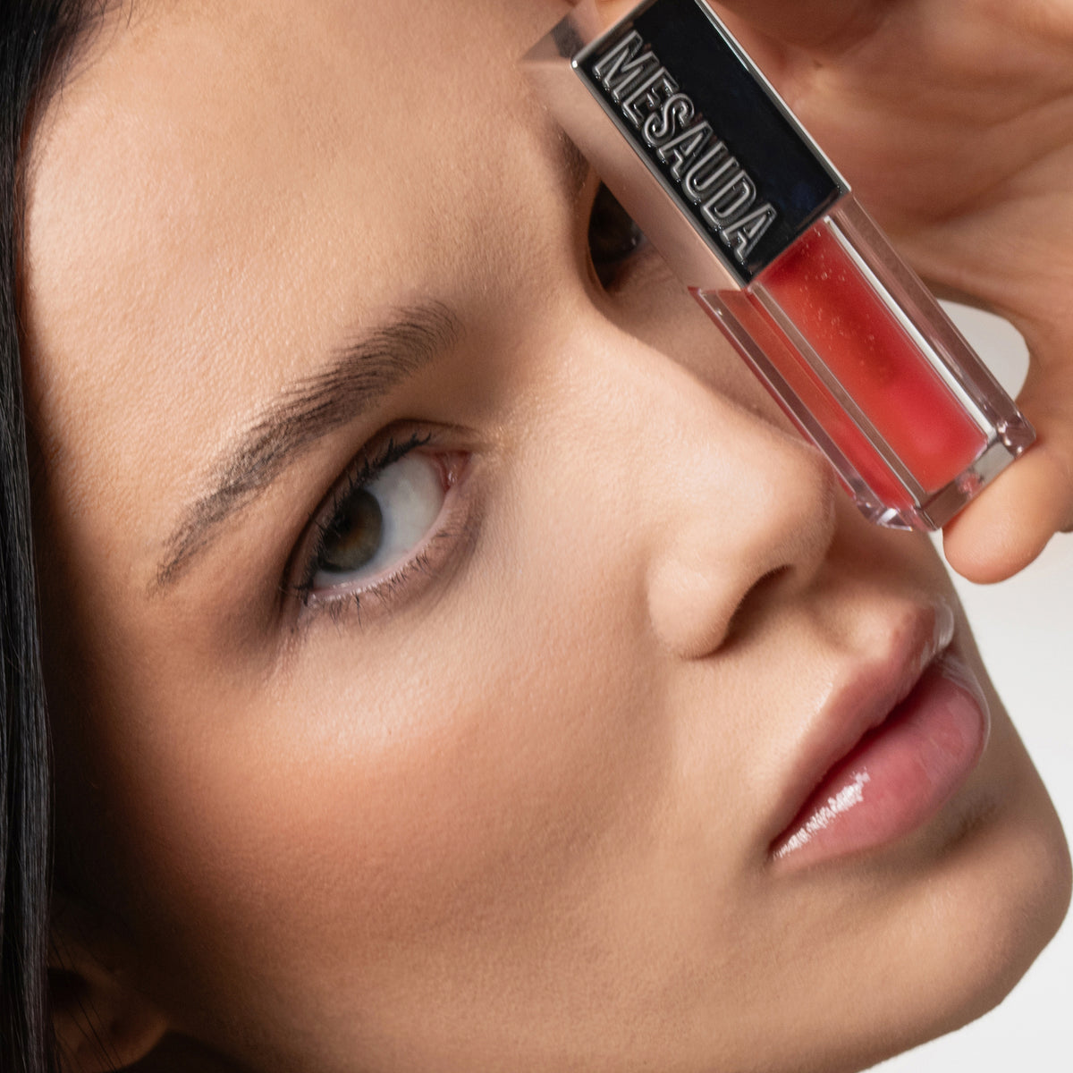 Eine Frau hält einen Mesauda Beauty LIPOILOGY™ Lipgloss in die Nähe ihres Auges, um ihre glänzenden Lippen zu betonen und blickt in die Kamera.