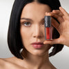Eine Frau mit glattem schwarzem Haar hält einen Mesauda Beauty LIPOILOGY™ Lipgloss in die Nähe ihres Gesichts, um ihre Lippen zu betonen.