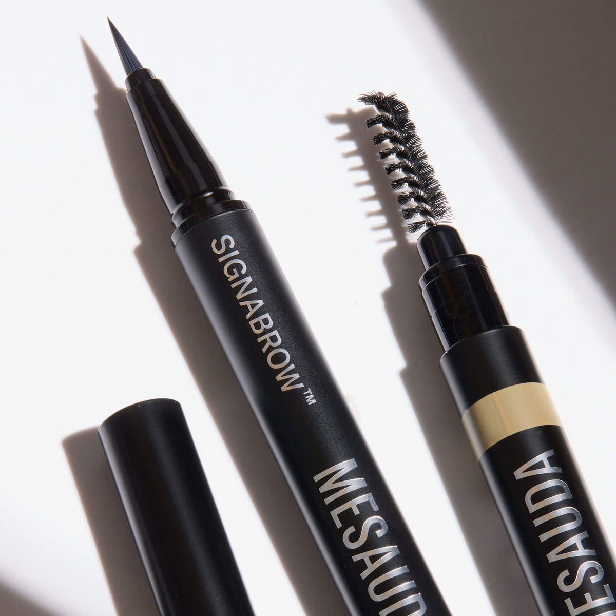 Zwei schwarze Augenbrauenstifte von Mesauda Beauty Signabrow - eine feine Spitze und eine Bürste - sind auf einem weißen Hintergrund mit Schatten abgebildet.