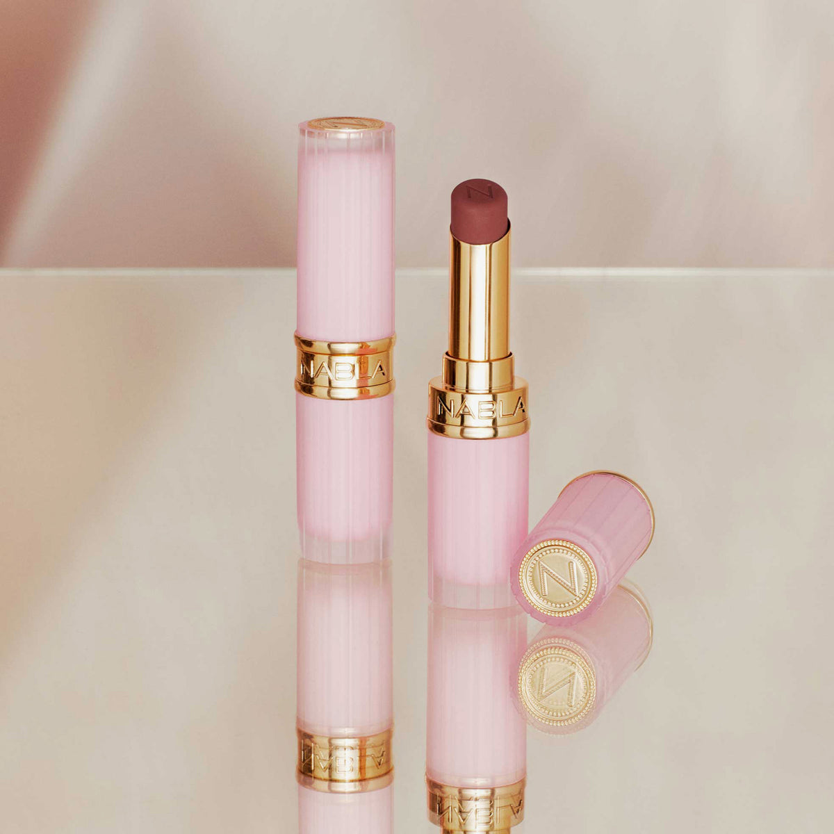 Zwei Beyond Blurry-Lippenstifte in Pink und Gold, einer offen und einer geschlossen, präsentiert auf einer reflektierenden Oberfläche.