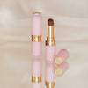 Zwei Beyond Blurry-Lippenstifte in Pink und Gold, einer offen und einer geschlossen, präsentiert auf einer reflektierenden Oberfläche.