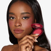 Eine Frau mit glatter Haut hält ein geöffnetes Close-Up Blurring Powder Blush, trägt den Blush auf den Wangenknochen und natürliches Make-up und blickt in die Kamera.