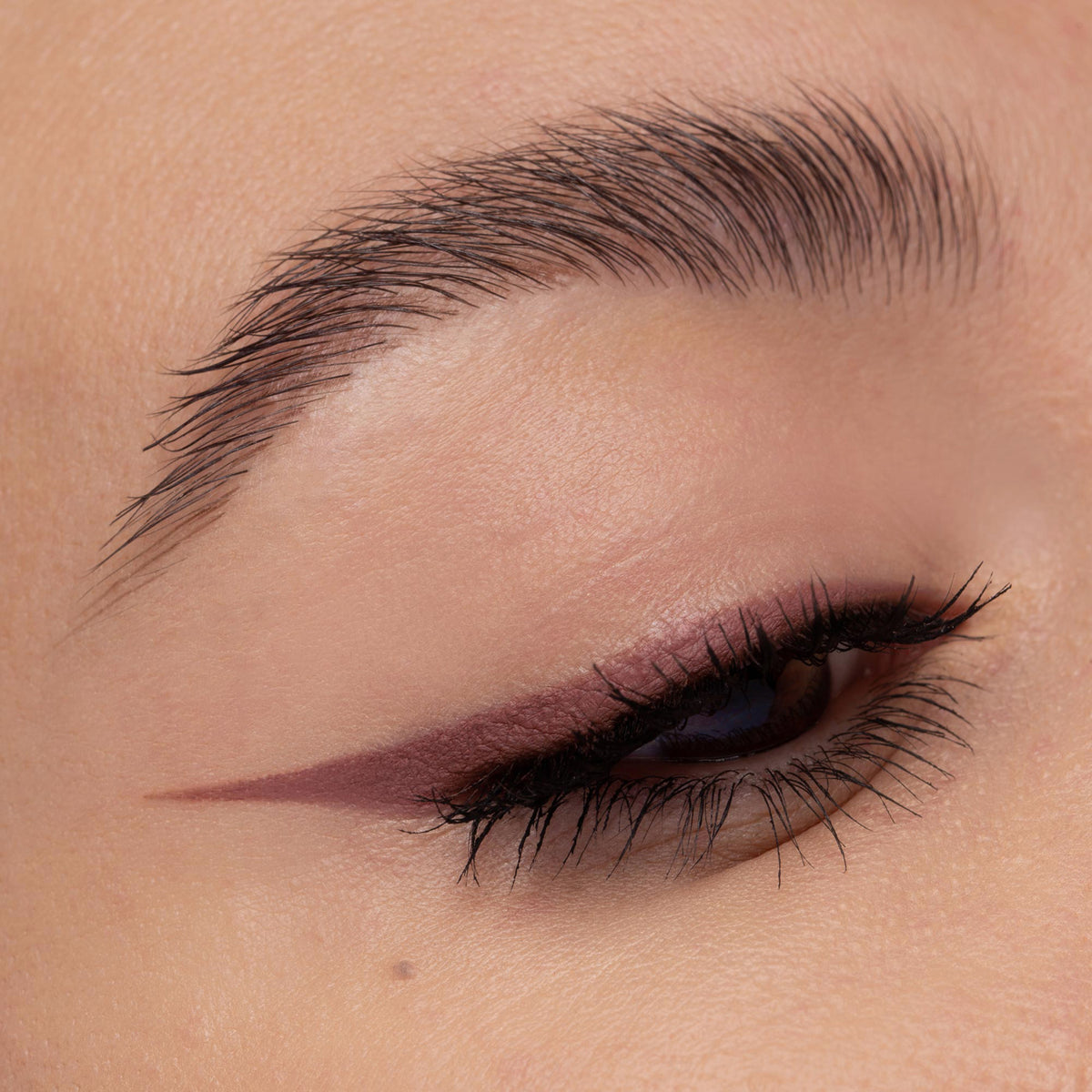 Geschminktes Auge mit rosébraunem Winged Eyeliner-Look, der sanft am äußeren Lidrand ausläuft. Die Wimpern sind schwarz und definiert.
