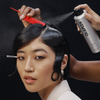 Hände, die das Haar einer Person mit einem Kamm und einer Dose OLAPLEX Shape Set Hairspray stylen.