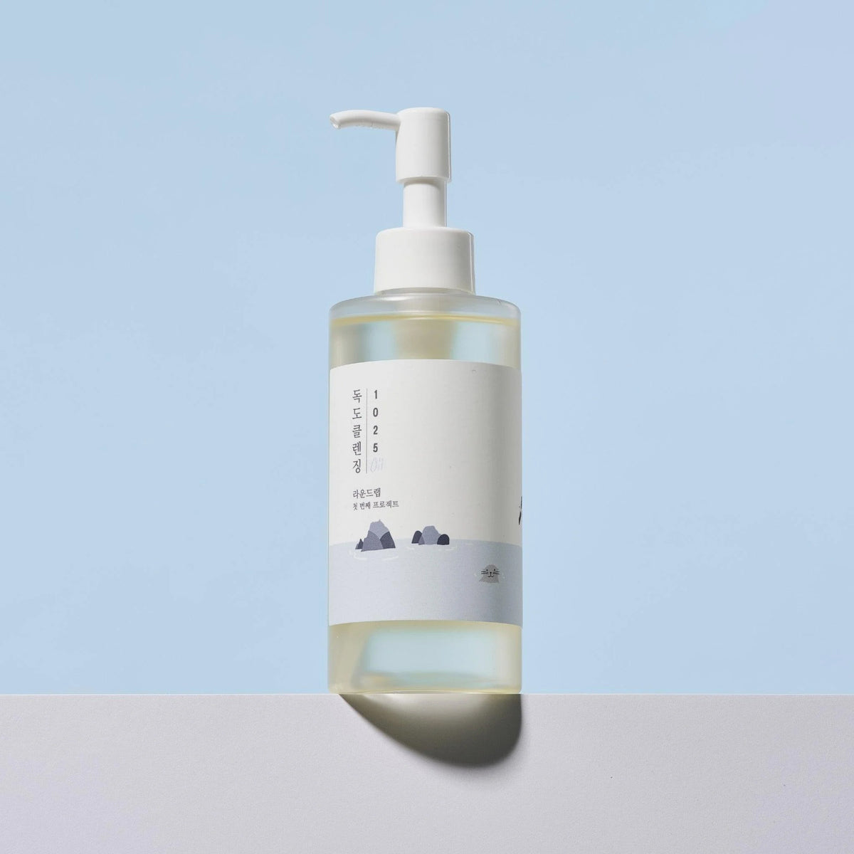 Mattiert transparente Flasche des 1025 Dokdo Cleansing Oil von Round Lab mit Pumpspender auf hellem Untergrund vor blauem Hintergrund.