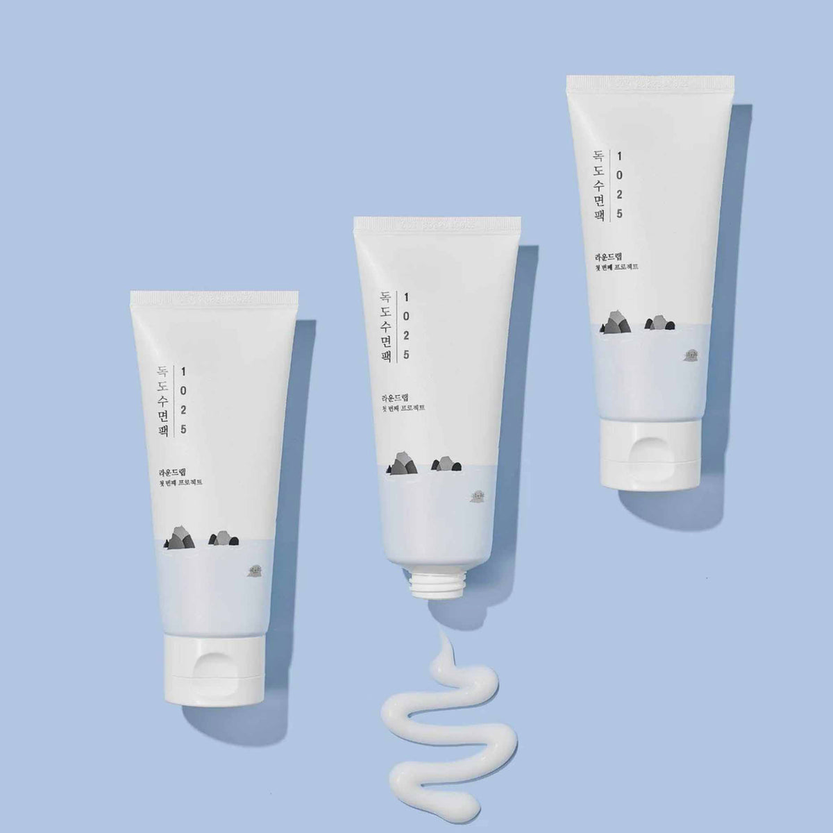 Drei Tuben des Round Lab 1025 Dokdo Sleeping Pack auf hellblauem Hintergrund, mittlere Tube geöffnet mit herausgedrückter, weißer Gel-Textur in geschwungener Form.