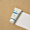 Eine Tube Birch Juice Moisturizing Cleanser liegt auf sandigem Boden mit einem weißen Streifen und zeigt auf der Verpackung eine Schwimmerillustration.