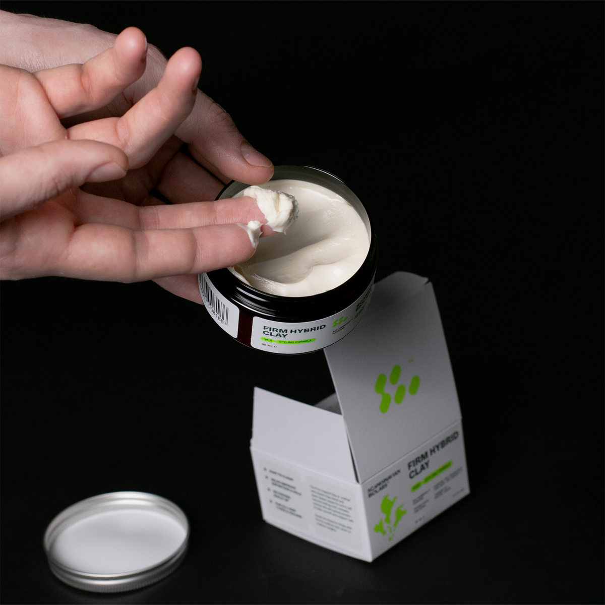 Eine Hand löffelt weißes Stylinggel aus einem Tiegel Scandinavian Biolabs Firm Hybrid Clay, in dessen Nähe sich die offene Schachtel befindet.