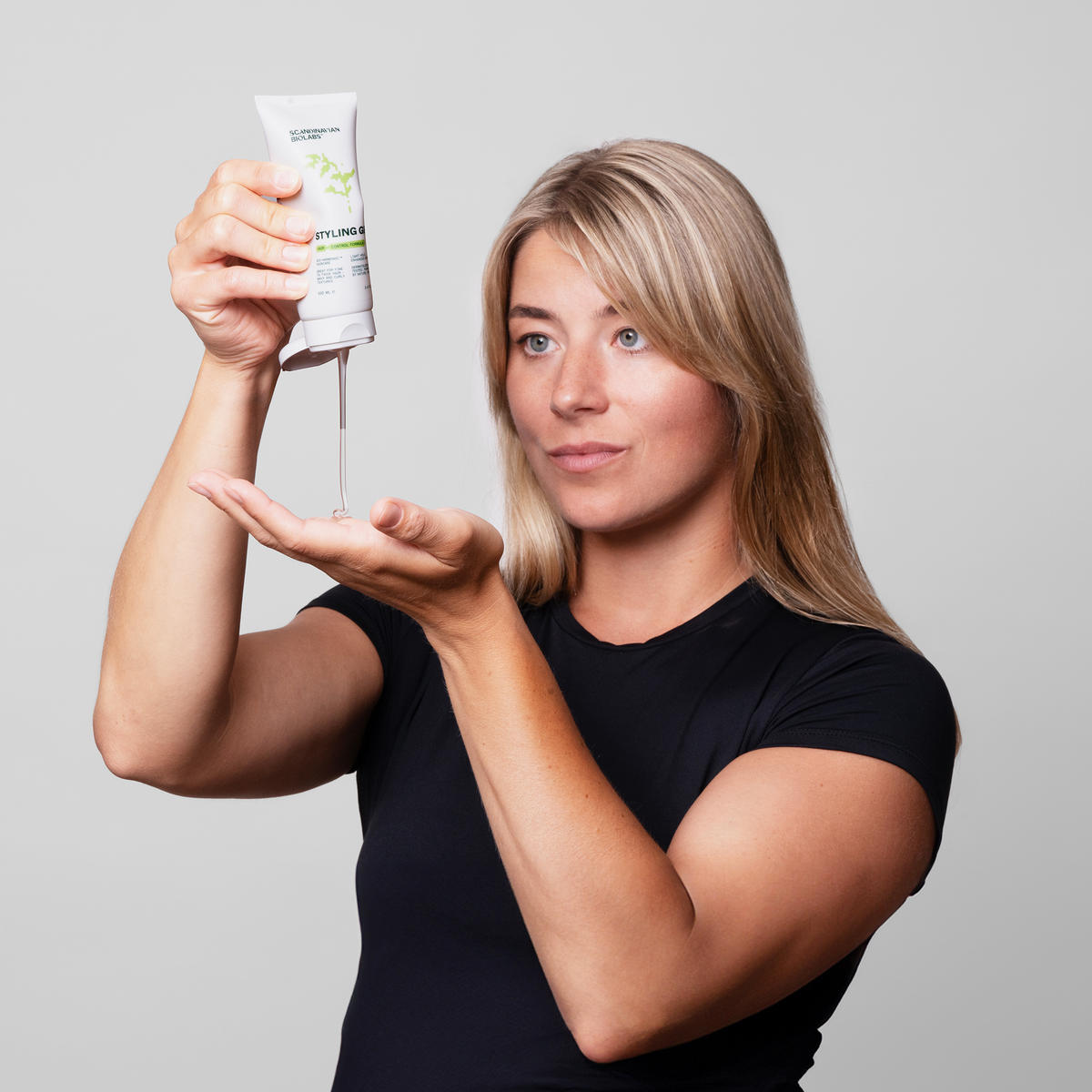 Eine Frau in einem schwarzen Hemd verteilt Scandinavian Biolabs Hair Styling Gel aus einer Tube in ihre Handfläche vor einem schlichten Hintergrund.