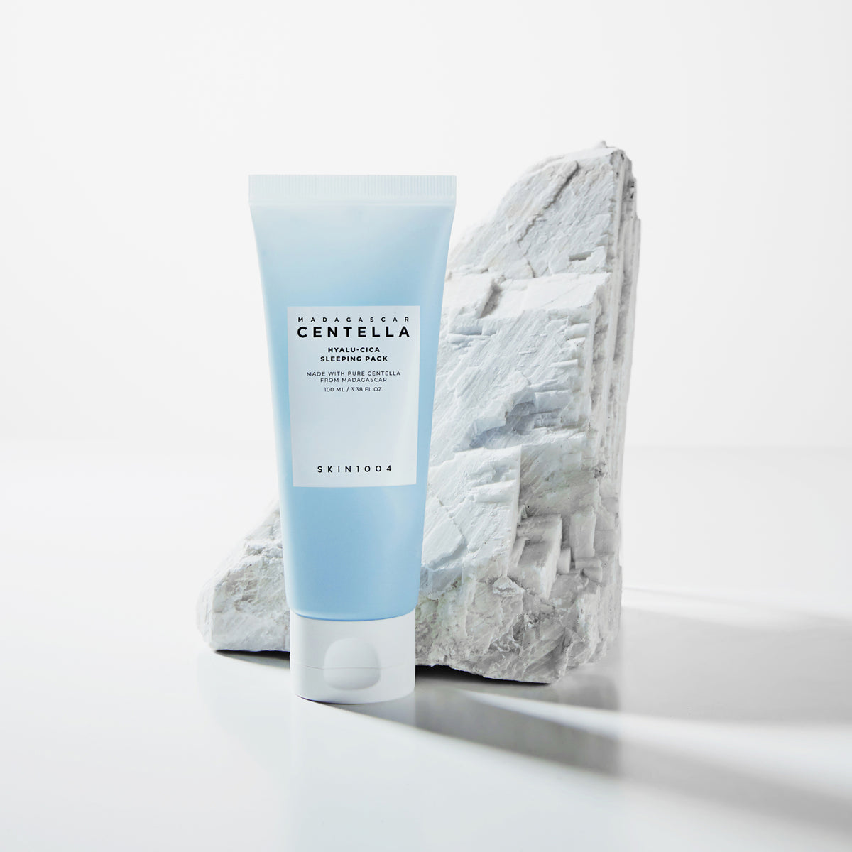 Eine blaue Tube Madagascar Centella Hyalu-Cica Sleeping Pack steht vor einem strukturierten weißen Stein auf weißem Hintergrund.