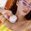Frau mit Sonnenbrille und gelbem Oberteil faulenzt im Freien und hält einen Tiegel Cloud Cream Light in der Hand.