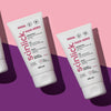 Drei Tuben Hair Hero-Creme werden auf einem rosa und violetten Hintergrund mit sichtbaren Schatten präsentiert.