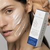 Frau mit aufgetragener Creme im Gesicht hält eine Skintegra Architect Serumverpackung in ihrer Hand.