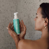 Frau in der Dusche hält eine nasse, türkisfarbene Flasche des Skintegra Clarion 2% BHA Cleansers in den Händen und betrachtet sie aufmerksam.
