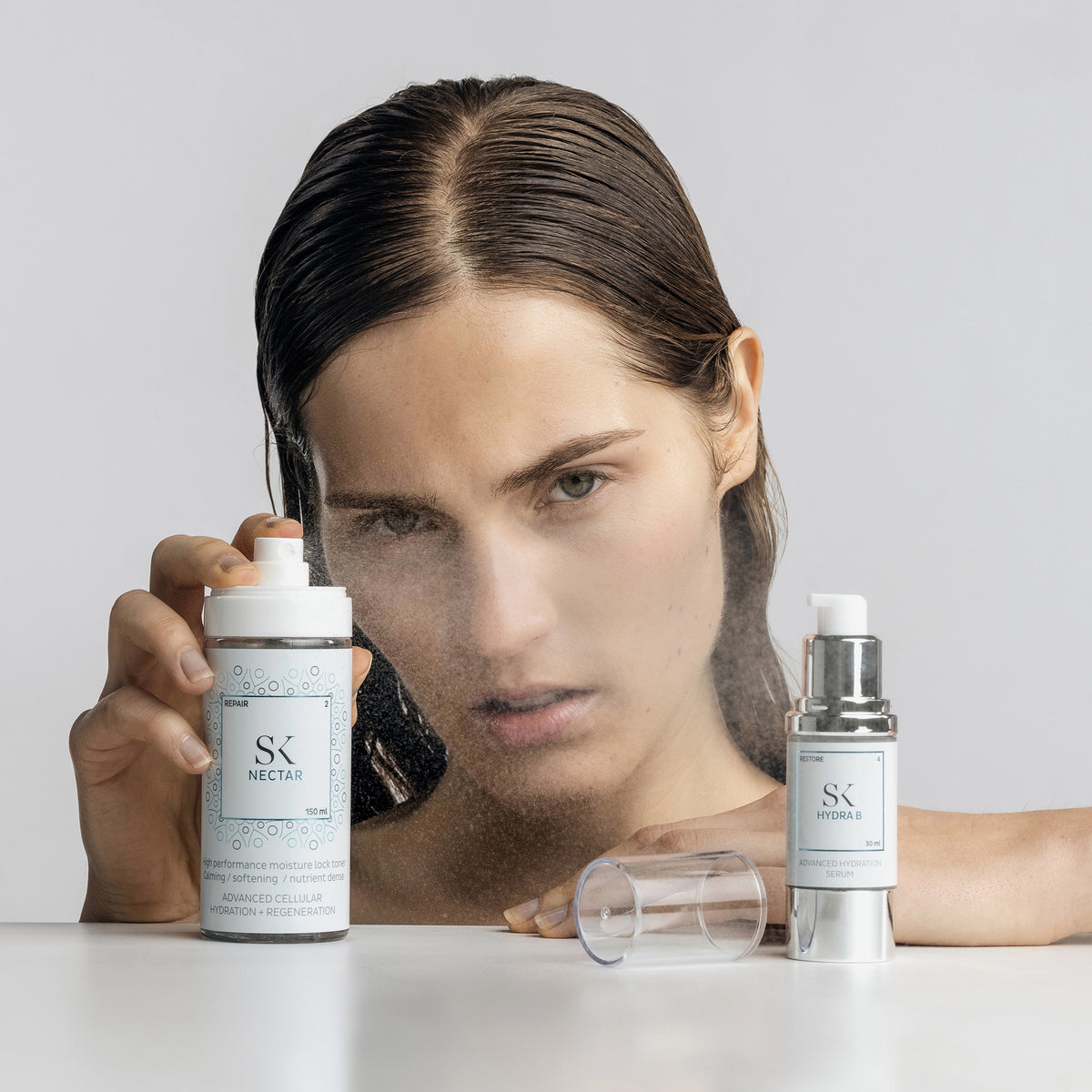 Frau mit nassen Haaren sprüht Skintegra Nectar Toner; daneben steht Skintegra Hydra B Serum auf weißem Tisch.