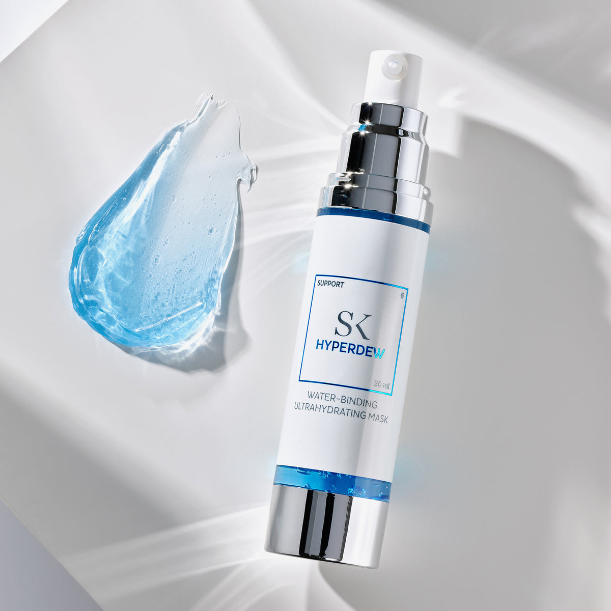 Weiße Flasche der Skintegra Hyperdew Maske mit silbernem Pumpspender, daneben ein Tropfen blauer Geltextur auf hellem, glänzendem Untergrund.