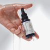Nasse Hand hält eine tropfende, violette Glasflasche mit schwarzer Pipette. Auf dem Etikett steht „SK Naro – Ultralight Oil Booster“.