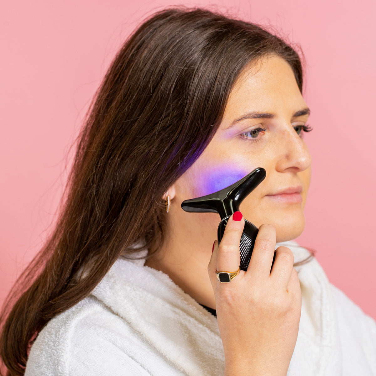 Frau nutzt ein schwarzes STYLPRO-Gerät mit blauem LED-Licht zur Gesichtsmassage auf der Wange, vor rosa Hintergrund.