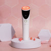 Facial Tool mit reinem roten LED-Licht auf rosafarbenem sechseckigem Hintergrund, platziert auf einem runden Ständer mit kleinen rosafarbenen Blumen.