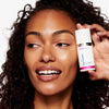 Eine Frau lächelt, während sie vor einem weißen Hintergrund eine Flasche Dark Circle Eye Cream nah an ihr Gesicht hält.