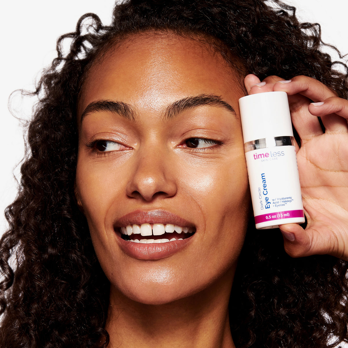 Eine Frau lächelt, während sie vor einem weißen Hintergrund eine Flasche Dark Circle Eye Cream nah an ihr Gesicht hält.