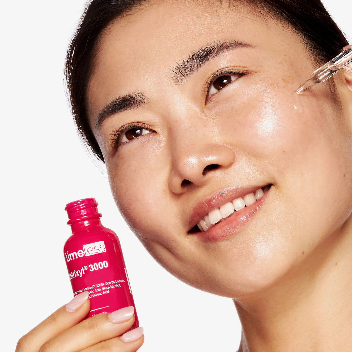 Eine Frau lächelt, während sie das Matrixyl 3000 Serum mit einer Pipette auf ihre Wange aufträgt und dabei die pinke Flasche hält.