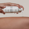Eine Person trägt AHA Smoothing Body Lotion aus einer weißen Flasche auf ihr Bein auf.