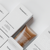 Eine Tube Bakuchiol Firming Cream in Braun liegt halb aus der Verpackung; umgeben von weiteren geschlossen Verpackungen.