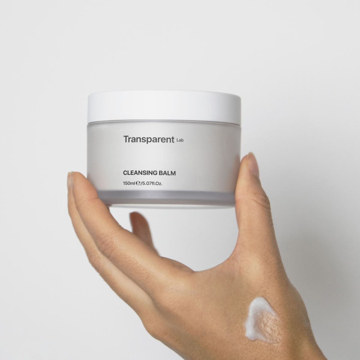 Eine Hand hält ein Glas Transparent Lab Cleansing Balm und trägt einen Teil des Balsams auf den Handrücken auf.
