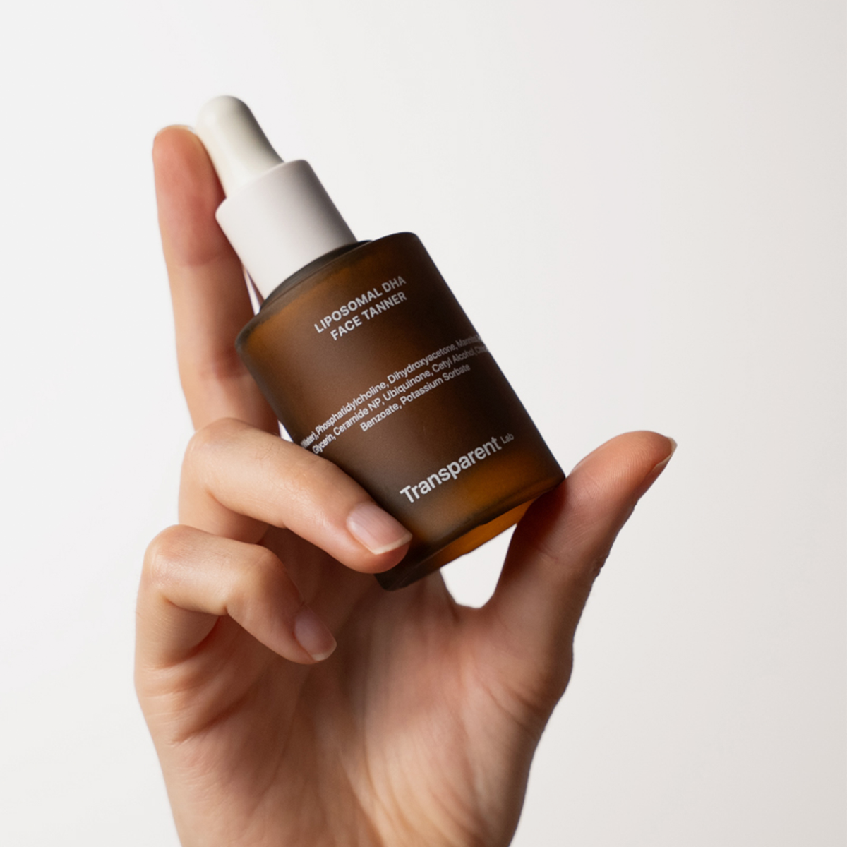 Eine Hand hält eine braune Tropfflasche mit der Aufschrift Liposomal DHA Face Tanner von Transparent Lab.