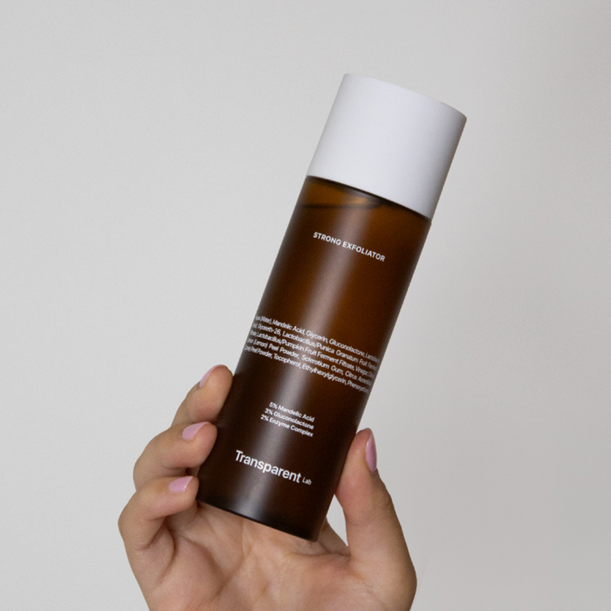Eine Hand hält eine braune Flasche Transparent Lab Strong Exfoliator mit einer weißen Kappe vor einem schlichten Hintergrund.