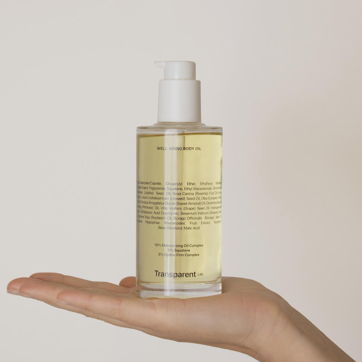 Eine klare Pumpflasche mit Well-Aging Body Oil von Transparent Lab wird auf einer offenen Hand vor einem schlichten Hintergrund gehalten.