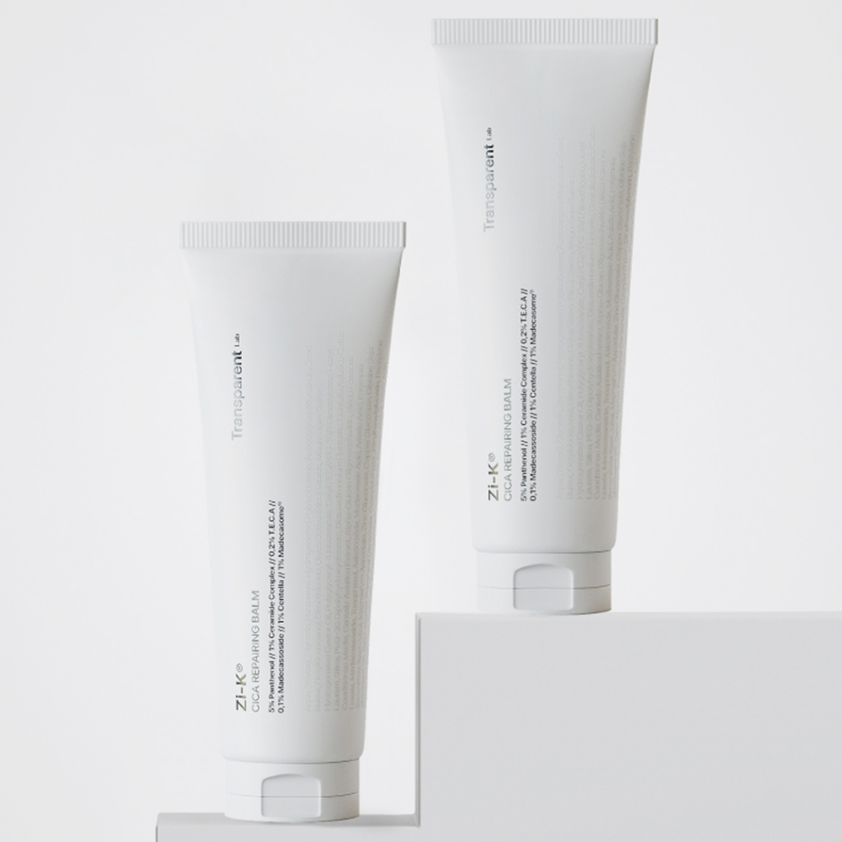 Zwei weiße Tuben Transparent Lab Zi-K® Cica Repairing Balm stehen aufrecht auf weißen Plattformen.
