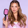Eine Frau mit langem, gewelltem Haar hält die UNbrush Detangling Hair Brush in Pink und trägt eine lavendelfarbene Strickjacke vor einem lila Hintergrund.