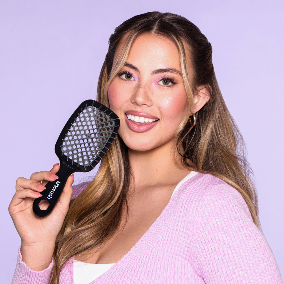 Eine Frau in einem rosafarbenen Oberteil lächelt, während sie die schwarze UNbrush Detangling Hair Brush vor einem leuchtend lila Hintergrund in der Hand hält.