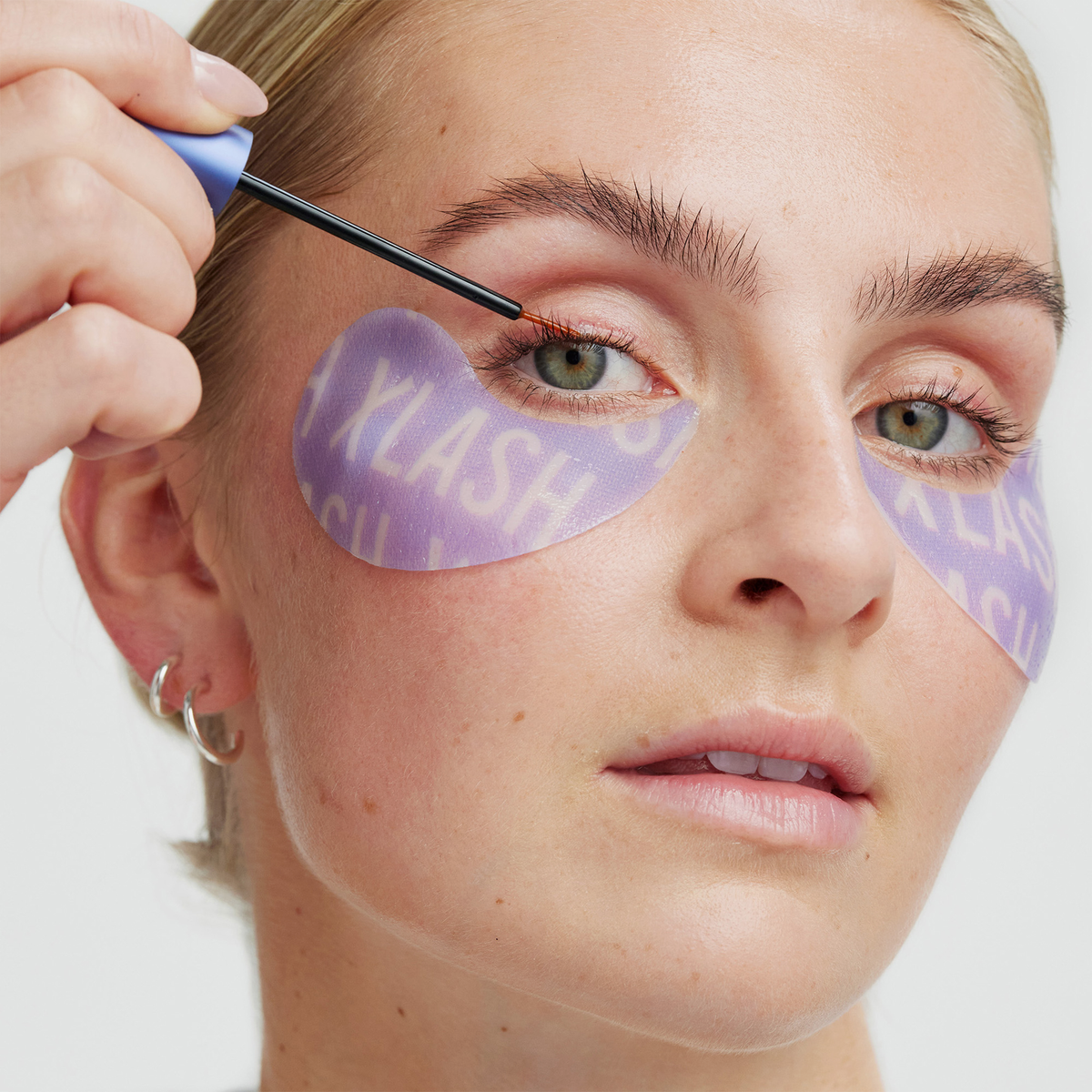 Eine Person trägt Mascara auf, während sie die XLASH Eyes Glow-up Holiday Kit-Pflaster mit weißer Aufschrift trägt und dabei geradeaus schaut.