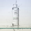 Eine Flasche Heartleaf 77 % Soothing Toner, um die herum Wasser auf einer klaren Oberfläche spritzt.