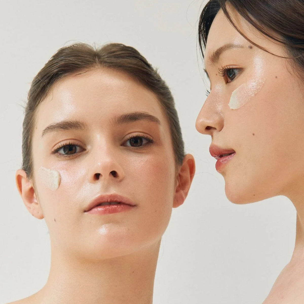 Zwei Frauen mit je einem Streifen Ground Rice and Honey Glow Mask auf einer Wange vor weißem Hintergrund.