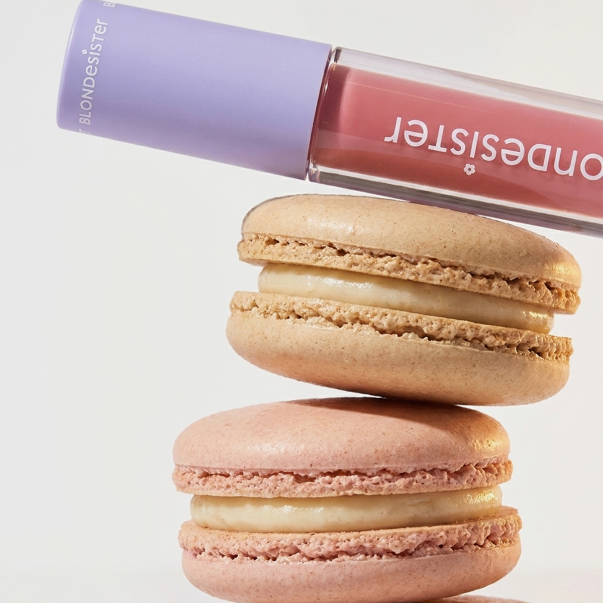BLONDESISTER 2 IN 1 SWEET GLOSS TINTED LIP OIL präsentiert sich auf einem Stapel aus rosa und beigen Macarons mit Sahne vor einem weißen Hintergrund.