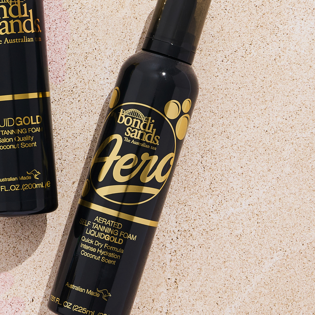 Zwei Flaschen Bondi Sands Aero Self Tanning Foam Liquid Gold auf Sand, von oben im hellen Sonnenlicht fotografiert.
