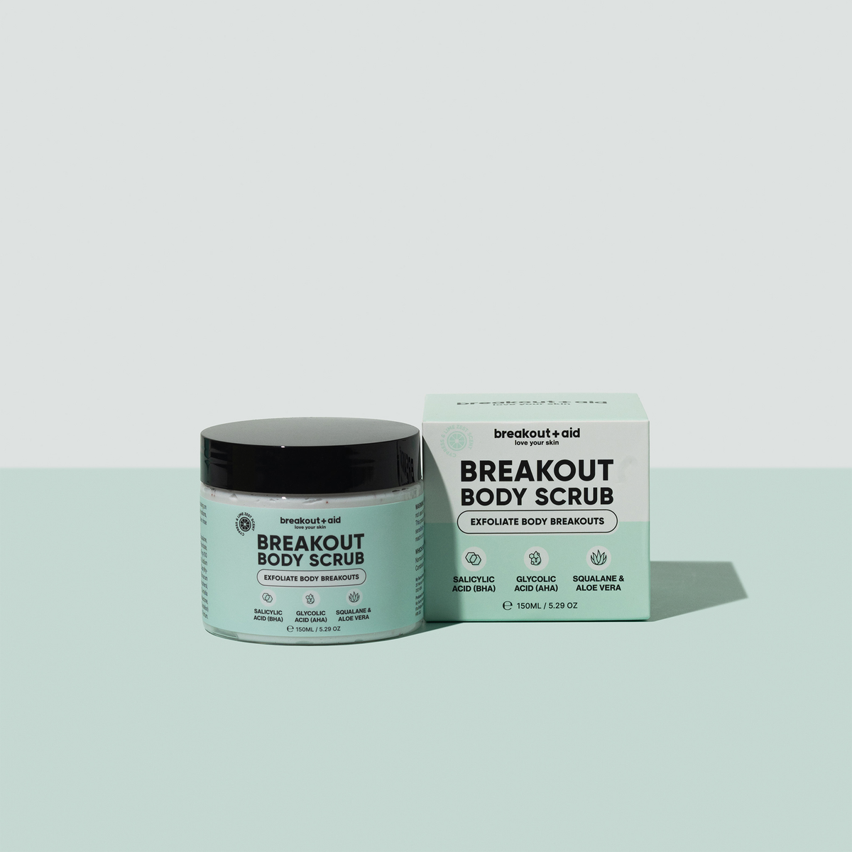 Ein Tiegel Breakout Body Scrub neben seiner quadratischen Verpackung; auf beiden sind auf den Etiketten Salicylsäure, Glykolsäure, Squalan und Aloe Vera aufgeführt.