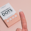 Ein Finger, der ein kleines, durchscheinendes, rundes Emergency Dots with Salicylic Acid-Akne-Pflaster hält, im Hintergrund eine Produktverpackung von Breakout+Aid.